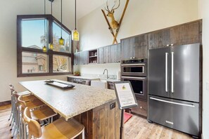 A modern kitchen with a large island, stainless steel appliances, and a view through a window. - Inviting condo with skiing, pool, fireplace, mountain view, & W/D (Sun Valley)