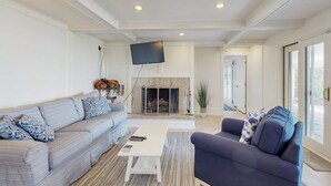 Un salón con sofá, sillón, chimenea, televisión y ventanas amplias.