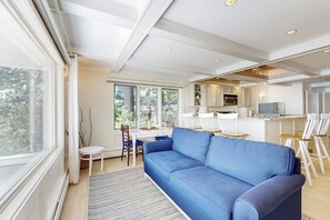 A modern living room with a blue sofa, a dining area with white chairs, and a kitchen with white cabinets.