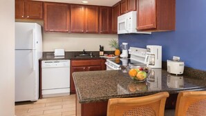 Une cuisine avec des meubles de rangement en bois, un réfrigérateur blanc, un lave-vaisselle, un four à micro-ondes, une machine à café et un panier de fruits posé sur le plan de travail.
