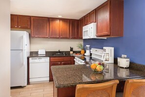 A kitchen with wooden cabinets, a white refrigerator, a dishwasher, a microwave, a coffee maker, and a fruit bowl on the counter. - Oceanfront, modern in Kailua-Kona. It’s ideal for all of your island adventures. (Kailua-Kona)