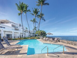 A pool area with lounge chairs, a clear blue sky, palm trees, and a view of the ocean. - Oceanfront, modern in Kailua-Kona. It’s ideal for all of your island adventures. (Kailua-Kona)