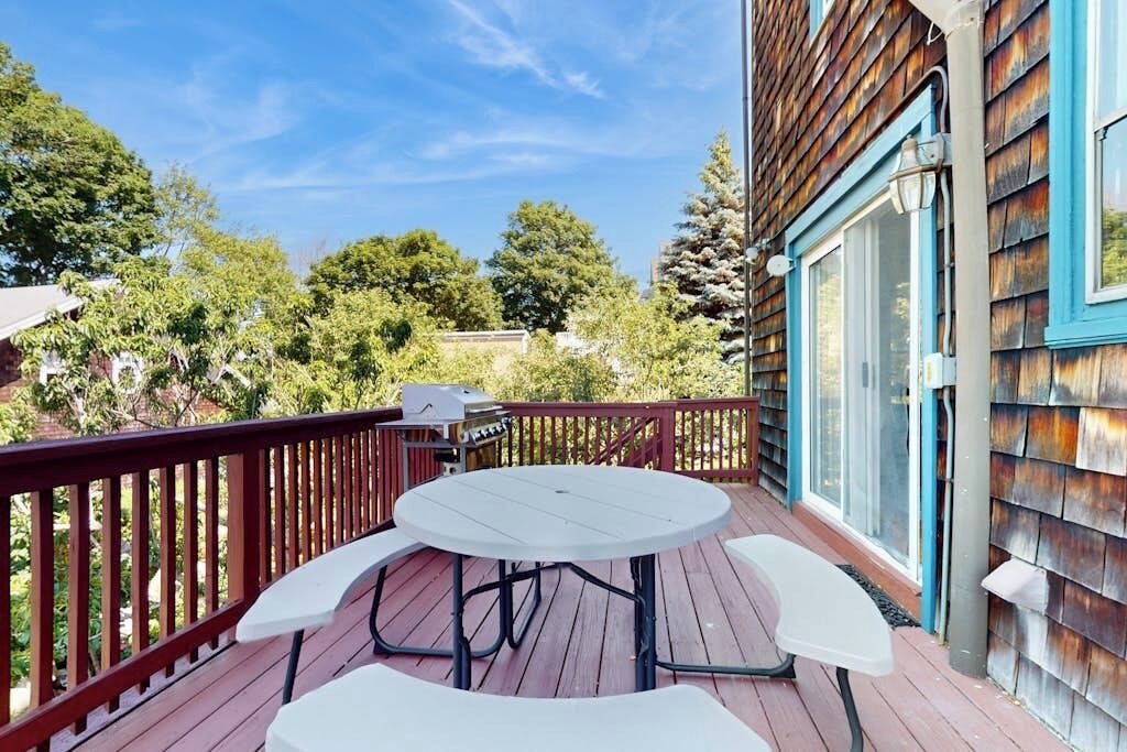 Une terrasse en bois avec une table ronde et des chaises, un barbecue et une vue sur des arbres et un bâtiment.