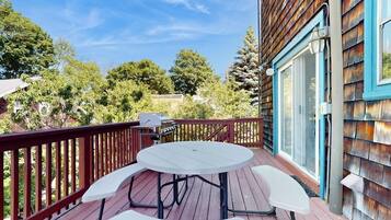 Une terrasse en bois avec une table ronde et des chaises, un barbecue et une vue sur des arbres et un bâtiment.