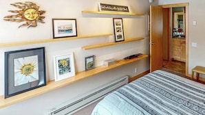 A bedroom with a bed, a wall-mounted shelf with framed pictures, a sun decoration, and a door leading to a bathroom. - Cozy ski condo with patio, fireplace, WiFi, shared pool & hot tub, walk to lifts (Sun Valley)
