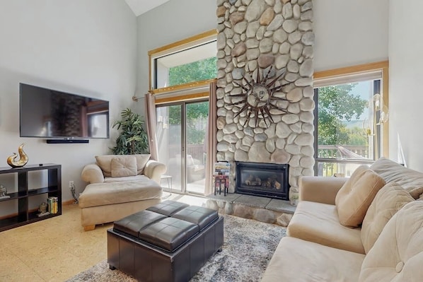 A living room with a fireplace, a flat-screen TV, a sofa, and a stone wall. - Golf Course Condo - dog friendly w/ community pool, hot tub & tennis courts (Sun Valley)