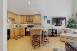 Cocina con armarios de madera, microondas y estufa. En la zona de estar hay dos taburetes de bar y un televisor.
