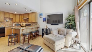 Une cuisine moderne avec des meubles en bois, un îlot central doté de tabourets, un téléviseur à écran plat fixé au mur et un coin salon avec un canapé et un pouf.