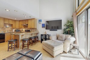 A modern kitchen with wooden cabinets, a central island with stools, a flat-screen TV mounted on the wall, and a seating area with a sofa and ottoman. - Golf Course Condo - dog friendly w/ community pool, hot tub & tennis courts (Sun Valley)
