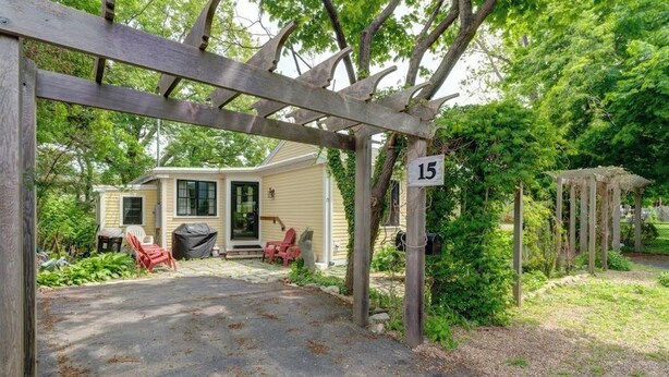 A covered driveway leading to a house with a number 15 sign, a covered porch, and a garden area.