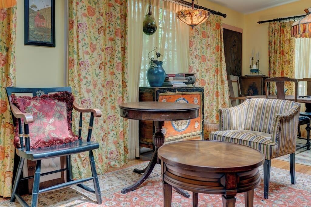 Une pièce avec un fauteuil ancien, une table ronde en bois et des rideaux à motifs floraux.