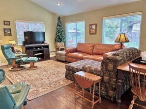 Ein Wohnzimmer mit Fernseher, Ledersofa, einem gemusterten Ottomanen, blauen Sesseln, einem Weihnachtsbaum und einem großen Fenster.