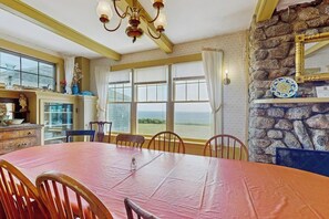 A dining area with a large table, chairs, a chandelier, a stone wall, and a view through the window.