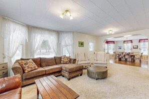 A spacious living room with a sectional sofa, a round ottoman, and a wooden coffee table. There is a dining area with a table and chairs in the background. - Charming Farmhouse on 100 Acres W/ Mountain Views, WiFi, Porch & Livestock (Columbia Falls)