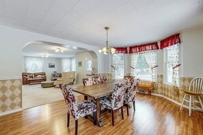 A dining area with a wooden table and patterned chairs, a chandelier, and a view of a living room with a sofa and a fireplace. - Charming Farmhouse on 100 Acres W/ Mountain Views, WiFi, Porch & Livestock (Columbia Falls)