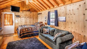 A cozy cabin with a wooden interior, a bed with a patterned blanket, a sofa with cushions, a ceiling fan, and a television on the wall. - Waterfront home on Echo Lake with dock, amazing view, deck & washer/dryer (Bigfork)