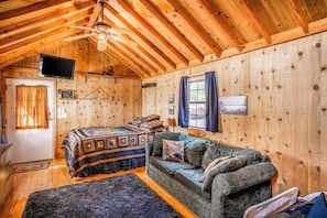 Ein gemütliches Blockhaus mit Holzvertäfelung, einem Bett mit gemustertem Überwurf, einer Couch mit Kissen, einem Deckenventilator und einem Fernseher an der Wand.