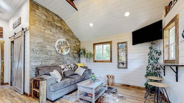 A rustic interior with a wooden wall, a grey sofa, a coffee table, and a TV mounted on the wall. - Rustic getaway with fireplace, patio area, board games, full kitchen, & TV (Kalispell)