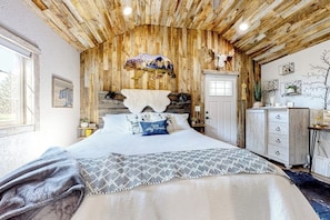 Ein rustikales Schlafzimmer mit Holzbalkendecke, einem Bett mit gemusterten Betttextilien, einem aufgehängten Tierkopf und einer Tür mit Fenster.