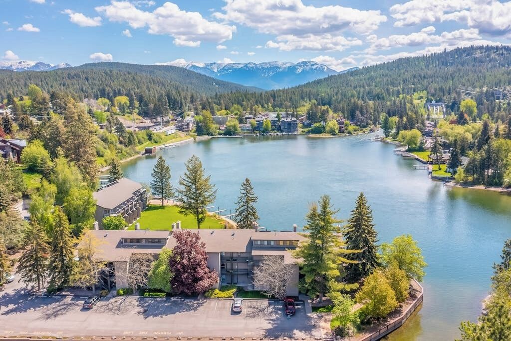 Vista de un lago con un edificio rodeado de árboles y una montaña al fondo.