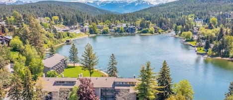 Vista de un lago con un edificio rodeado de árboles y una montaña al fondo.