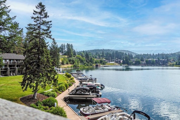 A lakeside view with docked boats, a building with a balcony, and a forested landscape. - Lakefront condo with balcony, stunning views, fast WiFi & W/D (Bigfork)