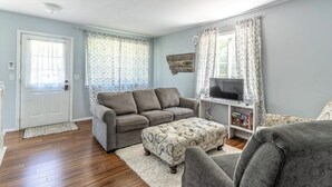 Un salon avec un canapé gris, un pouf à motifs, un meuble pour la télévision et une porte.