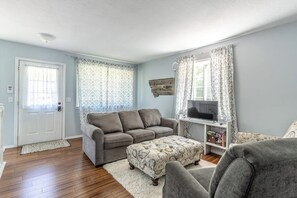 Ein Wohnzimmer mit grauem Sofa, einem gemusterten Hocker, einem Fernsehschrank und einer Tür.