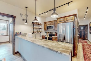 A modern kitchen with a marble countertop, stainless steel appliances, wooden cabinets, pendant lighting, and a 'Be Our Guest' wall decal. - Brand-new downtown condo with rooftop patio, grill, mtn views, fireplace, & W/D (Columbia Falls)