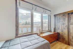 A bedroom with a large window, a bed, a wooden dresser, and a view of the outside. - Brand-new downtown condo with rooftop patio, grill, mtn views, fireplace, & W/D (Columbia Falls)