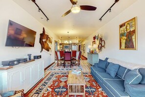A living room with a sofa, a coffee table, a TV, a ceiling fan, and a dining area with chairs and a table. - Brand-new downtown condo with rooftop patio, grill, mtn views, fireplace, & W/D (Columbia Falls)