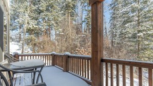 A wooden deck with a metal table and chairs, surrounded by snow-covered trees. - Amiable townhome with fireplace, hot tub, patio, gas grill, deck, & balcony (Columbia Falls)