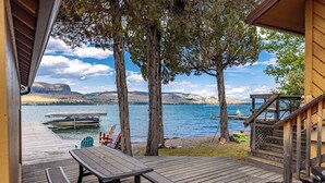 Una terraza de madera con una mesa de picnic, un barco amarrado al fondo y vista a un lago rodeado de montañas.