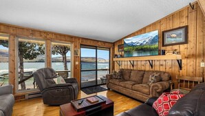 Una sala de estar con vista a un lago y montañas, amueblada con sofás de cuero y un televisor de pantalla plana.