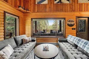 Ein Holzblockhaus-Interieur mit einem großen Fenster, zwei Sofas, einem runden Couchtisch und einem Schild mit der Aufschrift 'The Adventure Begins'.
