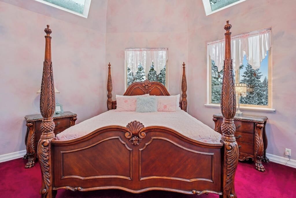 Een hemelbed met decoratieve houtsnijwerk, een groot raam met uitzicht op besneeuwde bomen, en een vloer bedekt met een rood tapijt.