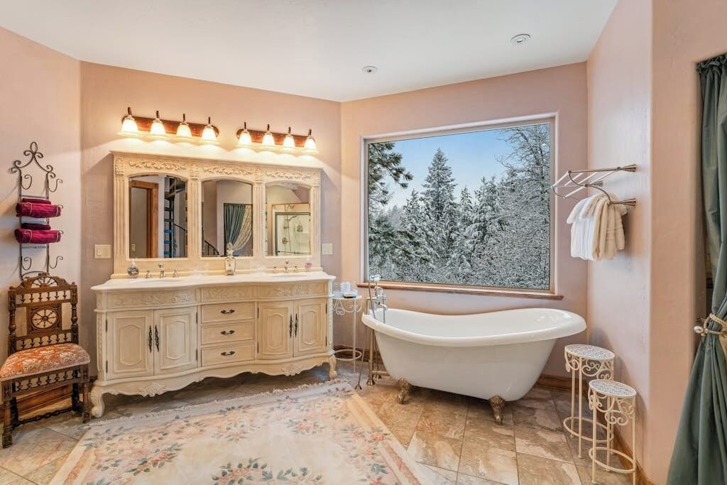 Une salle de bain avec une baignoire sur pieds, un meuble-lavabo doté d’un grand miroir et une vue sur des arbres couverts de neige.