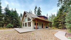 A modern house with a large porch, a covered outdoor seating area, and a gravel driveway. - +2-acre home with firepit, grill, & wood stove, kayaks, lawn games, & views (Bigfork)