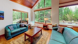 A spacious living room with a wooden ceiling, large windows, a blue sofa, a wooden coffee table, and a rug. - +2-acre home with firepit, grill, & wood stove, kayaks, lawn games, & views (Bigfork)