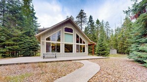 A lodge with a large windowed front, a covered porch, and a gravel driveway. - +2-acre home with firepit, grill, & wood stove, kayaks, lawn games, & views (Bigfork)