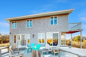 Ein zweistöckiges Haus mit einer Terrasse, großen Fenstern und einem überdachten Außenbereich zum Sitzen.
