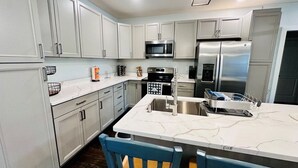A modern kitchen with white cabinets, stainless steel appliances, a marble countertop, and a bar area with two chairs. - Renovated Gulf Coast Home - Walk to Marina, Dining, & Downtown Carrabelle (Carrabelle)