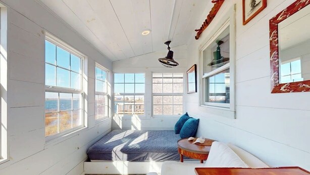 Una habitación con ventanas amplias, ventilador de techo, cama con almohadas azules y una mesa de madera.