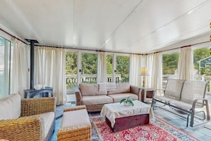 Ein sonnendurchflutetes Zimmer mit Korbmöbeln, einem roten Hocker und Blick auf viel Grün.