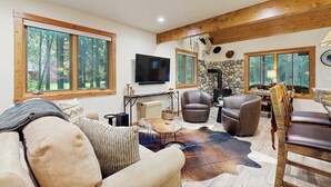A spacious living room with large windows, a stone fireplace, leather chairs, and a television. - Big Fork rustic loft cabin with forest views, WiFi, 5 miles to Flathead Lake (Bigfork)