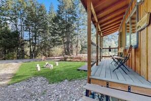 A wooden cabin with a porch, a grassy yard, and a forested backdrop. - Enjoy two separate cabins, pet friendly, firepit, 5 min to Lake, fly-fishing (Bigfork)