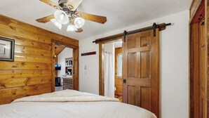 Un dormitorio con ventilador de techo de madera, una cama con cubrecama blanco, una puerta de madera y un pasillo que da a una zona de cocina.