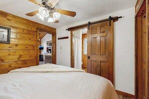 Ein Schlafzimmer mit Deckenventilator aus Holz, einem Bett mit weißer Bettdecke, einer Holztür und einem Gang, der in einen Küchenbereich führt.