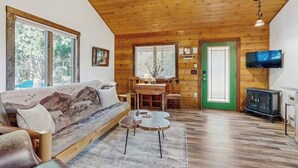 Interior acogedor de una cabaña con techo de madera, un sofá con estampado de paisaje, una mesa de comedor y un televisor.