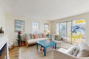 A living room with a sofa, a coffee table, and a chair. There are two windows and a view of the outside.